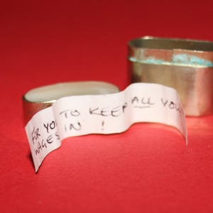 May include: A small, rectangular, tarnished metal box with a lid, next to a matching open box. A white paper strip with handwritten text that reads "FOR YOU TO KEEP ALL YOUR WAGES IN!" is draped over the open box.