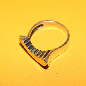 Peut inclure: Bague en argent au design unique. La bague pr&eacute;sente une face rectangulaire plate avec une s&eacute;rie de rainures parall&egrave;les. L'anneau est lisse et arrondi, cr&eacute;ant une esth&eacute;tique moderne et g&eacute;om&eacute;trique.