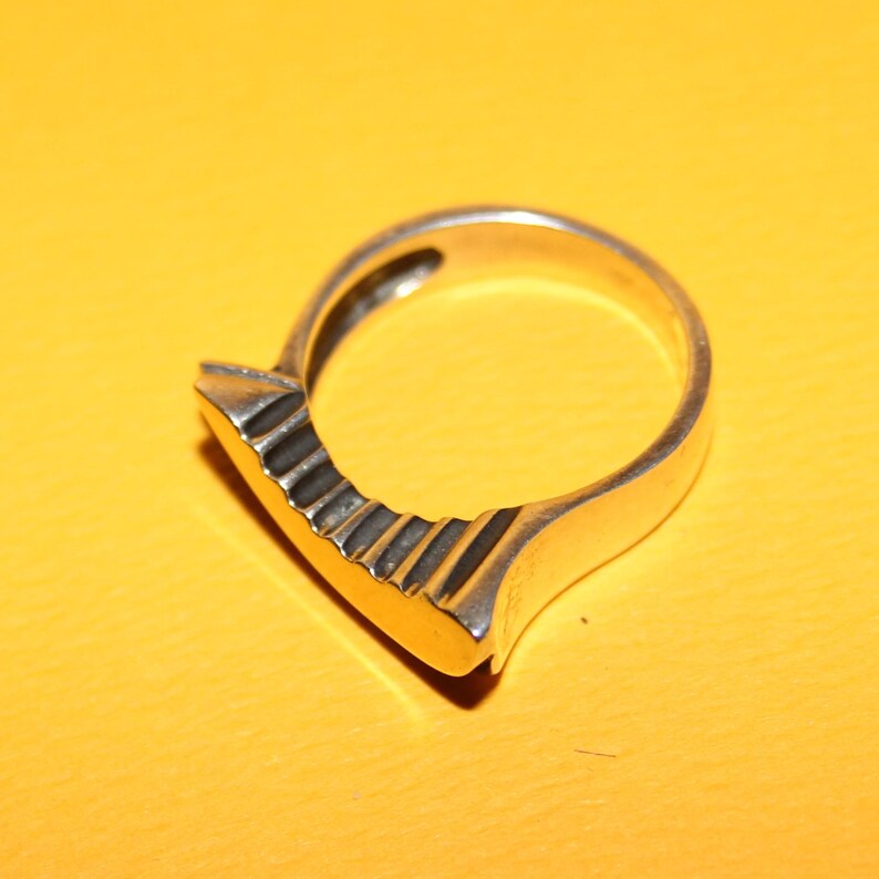 Peut inclure: Bague en argent au design unique. La bague pr&eacute;sente une face rectangulaire plate avec une s&eacute;rie de rainures parall&egrave;les. L'anneau est lisse et arrondi, cr&eacute;ant une esth&eacute;tique moderne et g&eacute;om&eacute;trique.