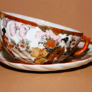 Peut inclure: Une tasse à thé et une soucoupe en porcelaine blanche avec une bordure rouge, orange et dorée. La tasse et la soucoupe sont décorées d'un motif floral et d'images de personnes en vêtements traditionnels japonais.