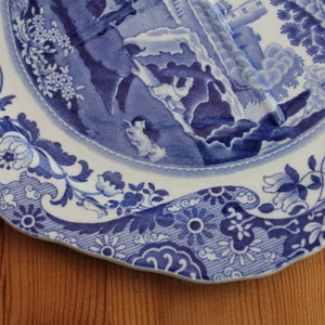 May include: A white and blue decorative plate with a detailed landscape scene in the center. The plate features a scalloped edge and floral patterns around the rim. The plate is resting on a wooden surface.