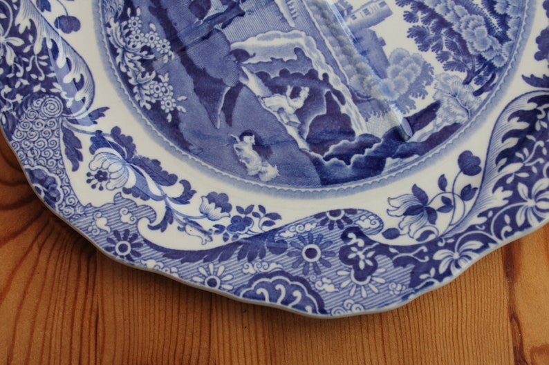 May include: A white and blue decorative plate with a detailed landscape scene in the center. The plate features a scalloped edge and floral patterns around the rim. The plate is resting on a wooden surface.