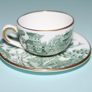 Può includere: Una tazza e un piattino bianchi con un design verde e oro. La tazza e il piattino presentano un motivo paesaggistico dettagliato con bordi dorati. La tazza ha un piccolo manico con accenti dorati. Ideale per servire il tè.
