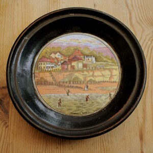 Könnte beinhalten: Runder, gerahmter dekorativer Teller mit einer gemalten Küstenlandschaft. Das Kunstwerk zeigt ein Dorf auf einer Klippe, mit Figuren, die im Wasser angeln. Der Teller ist in einem dunklen Holzrahmen gerahmt. Der Text "Pegwell Bay" ist sichtbar.