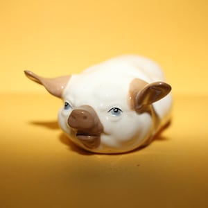 以下が含まれることがあります： 白い体と耳と鼻の茶色のアクセントが特徴のセラミック製の豚の置物。豚は青い目を持ち、横たわっています。置物は黄色の背景に配置されています。