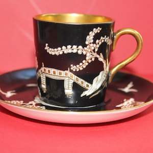 May include: Black teacup and saucer set with gold trim. The cup and saucer feature a detailed design of a bridge, birds, and floral elements in white and gold. The cup's interior is gold.