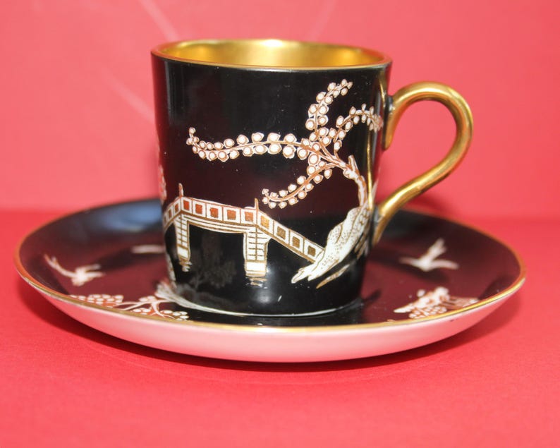 May include: Black teacup and saucer set with gold trim. The cup and saucer feature a detailed design of a bridge, birds, and floral elements in white and gold. The cup's interior is gold.