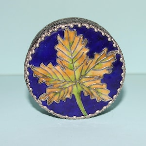 May include: A round, silver-toned trinket box with a vibrant design. The lid features a detailed yellow and green leaf against a deep blue background. The box has a decorative silver border.