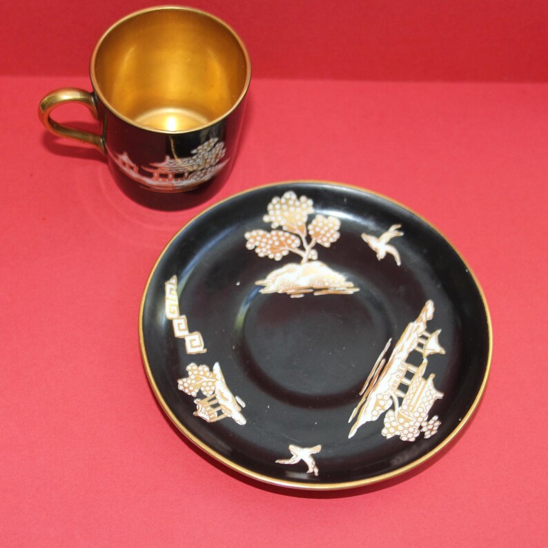May include: A black teacup and saucer set with gold trim. The cup and saucer feature gold and white Asian-inspired designs. The interior of the cup is gold. The saucer has a tree and bird design.