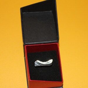 Peut inclure: Une bague argent&eacute;e au design incurv&eacute;, pr&eacute;sent&eacute;e dans un &eacute;crin noir et rouge. La bague pr&eacute;sente une surface textur&eacute;e avec des rayures argent&eacute;es et gris fonc&eacute; altern&eacute;es. L'&eacute;crin est ouvert, r&eacute;v&eacute;lant la bague sur un int&eacute;rieur en velours noir.