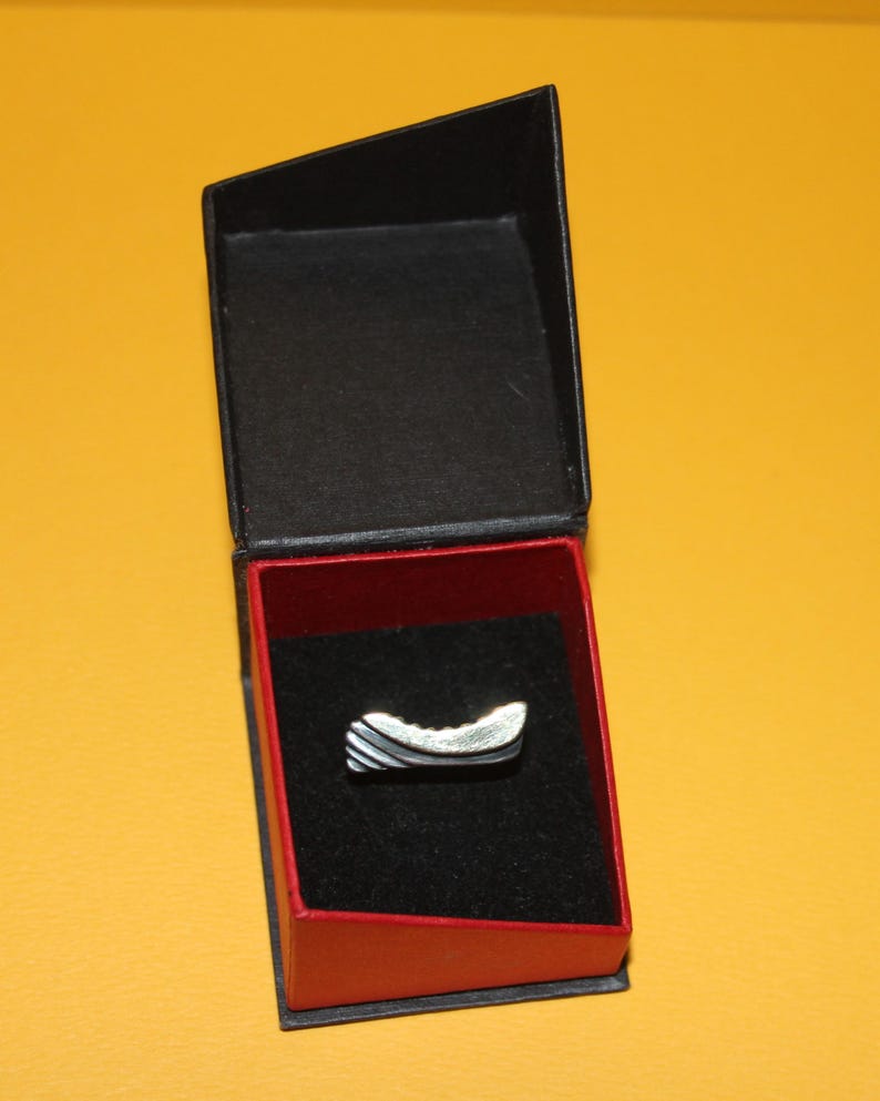Peut inclure: Une bague argent&eacute;e au design incurv&eacute;, pr&eacute;sent&eacute;e dans un &eacute;crin noir et rouge. La bague pr&eacute;sente une surface textur&eacute;e avec des rayures argent&eacute;es et gris fonc&eacute; altern&eacute;es. L'&eacute;crin est ouvert, r&eacute;v&eacute;lant la bague sur un int&eacute;rieur en velours noir.