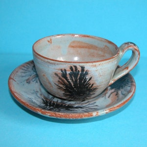 Può includere: Un set di tazza e piattino in ceramica fatto a mano. La tazza e il piattino sono azzurri con accenti marroni e neri. La tazza presenta un manico e un disegno nero a forma di felce. Il bordo della tazza e del piattino è marrone.