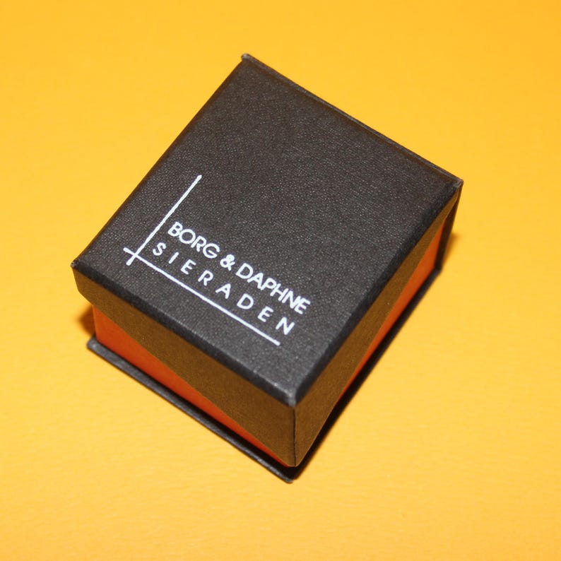 Peut inclure: Une petite bo&icirc;te noire rectangulaire avec le texte "BORG & DAPHNE SIERADEN" en blanc. L'int&eacute;rieur de la bo&icirc;te est orange. La bo&icirc;te est sur un fond jaune.