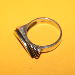 Peut inclure: Une bague en argent avec une face rectangulaire et un design en couches. La bague est pos&eacute;e sur un fond jaune, soulignant son &eacute;clat m&eacute;tallique et sa forme g&eacute;om&eacute;trique. L'anneau de la bague est lisse et arrondi.