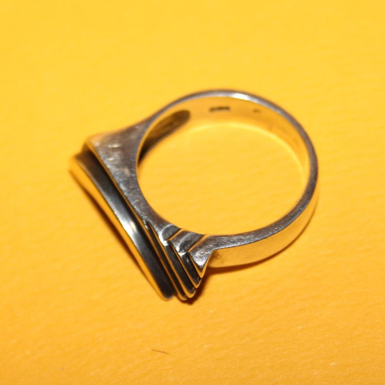 Peut inclure: Une bague en argent avec une face rectangulaire et un design en couches. La bague est pos&eacute;e sur un fond jaune, soulignant son &eacute;clat m&eacute;tallique et sa forme g&eacute;om&eacute;trique. L'anneau de la bague est lisse et arrondi.