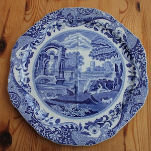 May include: A blue and white decorative plate with a landscape scene. The plate has a scalloped edge and floral border. The central image depicts a classical landscape with buildings, trees, and figures. The plate is set on a wooden surface.