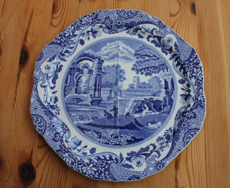 May include: A blue and white decorative plate with a landscape scene. The plate has a scalloped edge and floral border. The central image depicts a classical landscape with buildings, trees, and figures. The plate is set on a wooden surface.