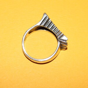Peut inclure: Une bague en argent au design g&eacute;om&eacute;trique unique. La bague pr&eacute;sente un dessus angulaire textur&eacute; avec une s&eacute;rie de lignes parall&egrave;les, cr&eacute;ant un look moderne et artistique. La bague est pos&eacute;e sur un fond jaune.