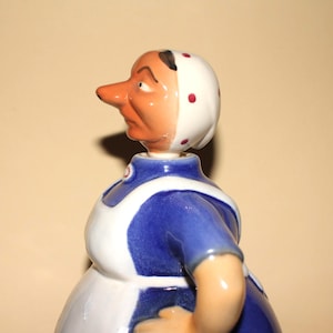 May include: A ceramic figurine of a woman in a blue and white uniform, with a white bonnet adorned with red polka dots. The figurine has a long nose and a stern expression. The figure is a vintage collectible.