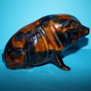 May include: A ceramic pig ornament with a glossy finish. The pig is brown and blue with a mottled pattern. The text "From Mother 1928" is inscribed on the side. The pig is posed in a standing position.