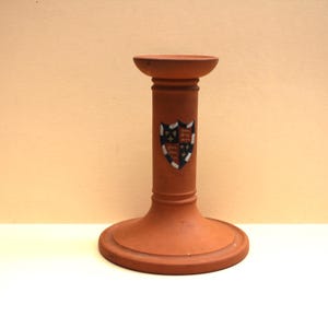 May include: A terracotta-colored candle holder with a cylindrical stem and a wide, circular base. The center of the stem features a shield-shaped emblem divided into four quadrants, displaying alternating red lions and gold fleur-de-lis symbols.