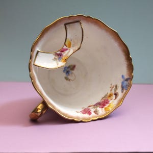 Könnte beinhalten: Eine weiße Porzellan-Schnurrbarttasse mit goldfarbenem, gewelltem Rand und integriertem Keramik-Schnurrbartschutz. Das Innere ist mit handgemalten Blumenmotiven in Rosa, Gelb und Blau verziert und verfügt über einen goldenen Henkel.