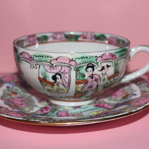 May include: A porcelain teacup and saucer set. The white cup and saucer feature a hand-painted design with figures in a garden scene, accented with pink, green, and gold trim. The set is placed on a pink background.