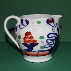 Puede incluir: Una jarra de cerámica blanca con un diseño floral azul, verde y rojo. La jarra tiene un cuerpo redondeado y un asa.