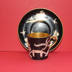 May include: A black teacup and saucer set with gold trim and Asian-inspired designs. The teacup and saucer have a dark background with gold and white accents. The teacup has a gold interior and handle.