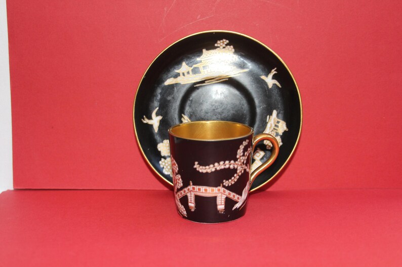 May include: A black teacup and saucer set with gold trim and Asian-inspired designs. The teacup and saucer have a dark background with gold and white accents. The teacup has a gold interior and handle.