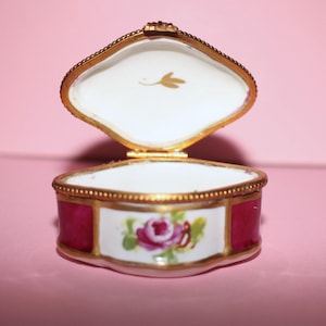 May include: An open porcelain trinket box with a hinged gold-tone metal rim. The exterior features alternating panels of deep magenta and white with a hand-painted pink rose motif. The interior lid displays a small gold leaf illustration on white.