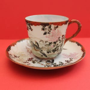 May include: A white teacup and saucer set with a detailed floral and bird design. The cup has a bamboo-style handle and a brown rim. The saucer and cup feature pink flowers and birds.