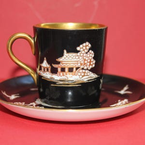 May include: A black teacup and saucer set with gold detailing. The cup has a gold handle and a white and gold Asian-inspired landscape design. The saucer has a white rim and matching design.