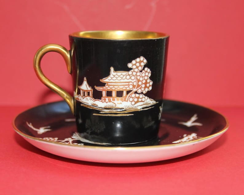 May include: A black teacup and saucer set with gold detailing. The cup has a gold handle and a white and gold Asian-inspired landscape design. The saucer has a white rim and matching design.