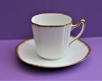 Puede incluir: Una taza de té y un platillo de porcelana blanca con un borde dorado. La taza tiene forma cuadrada y un asa.