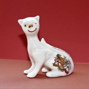 May include: White ceramic figurine of a Cheshire Cat winking with a green eye. The cat is sitting with its tail curled around its body. The cat has a coat of arms on its back with the text "CHESHIRE CAT. ST. WERBURGH CHESTER".