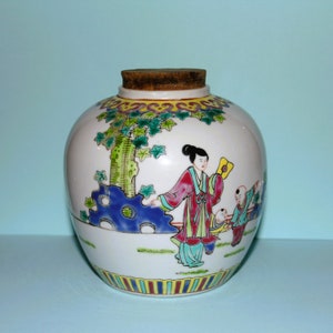 Puede incluir: Un tarro de porcelana blanca con tapa de corcho, decorado con una escena colorida de una mujer y dos niños en un jardín. El tarro tiene un patrón floral azul y blanco alrededor de la base.