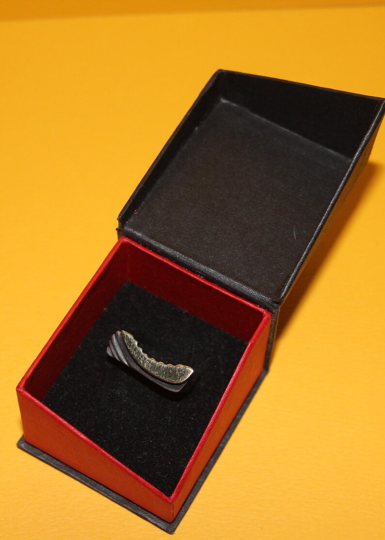 Peut inclure: Une petite bague incurv&eacute;e avec un design argent&eacute; et dor&eacute; repose &agrave; l'int&eacute;rieur d'une bo&icirc;te noire ouverte avec un int&eacute;rieur rouge. La bague a une surface textur&eacute;e et est expos&eacute;e sur une surface de velours noir. La bo&icirc;te est sur un fond jaune.