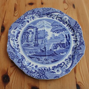 May include: A blue and white decorative plate with a landscape scene in the center. The plate has a scalloped edge and floral patterns. The landscape features buildings, trees, and figures. A classic piece for home decor.