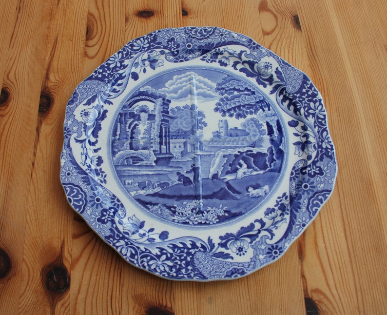May include: A blue and white decorative plate with a landscape scene in the center. The plate has a scalloped edge and floral patterns. The landscape features buildings, trees, and figures. A classic piece for home decor.