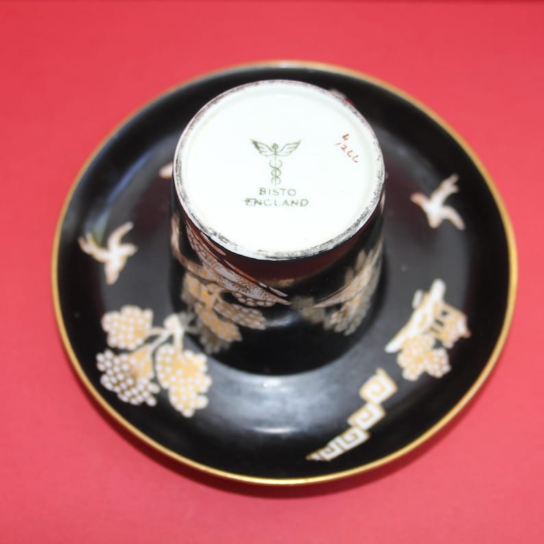 May include: A black teacup and saucer set with gold detailing. The cup and saucer are decorated with a floral and bird pattern. The edges of the saucer and cup are gold. The base of the cup reads "Bisto England".