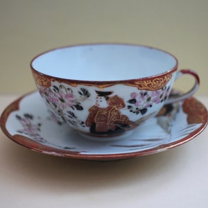 Peut inclure: Une tasse à thé et une soucoupe en porcelaine blanche avec un motif floral rouge et or. La tasse présente une image peinte d'une personne vêtue de vêtements traditionnels japonais.