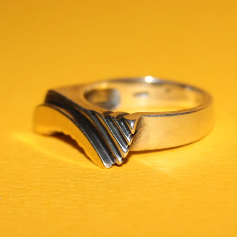 Peut inclure: Une bague en argent au design unique. La bague pr&eacute;sente une forme carr&eacute;e avec un accent incurv&eacute; et textur&eacute; sur un c&ocirc;t&eacute;. La zone textur&eacute;e comporte une s&eacute;rie de lignes parall&egrave;les, ajoutant un int&eacute;r&ecirc;t visuel &agrave; la pi&egrave;ce. La bague est pos&eacute;e sur un fond jaune.