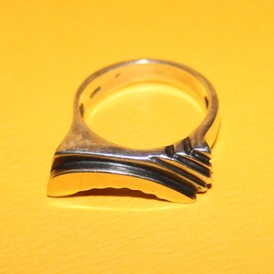 May include: A silver-toned ring with a unique design. The ring features a rectangular face with a series of horizontal lines and a curved bottom edge. The band is smooth and polished, set against a yellow background.
