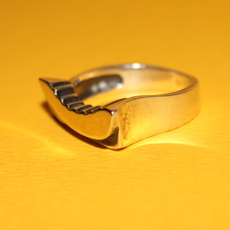 Peut inclure: Une bague en argent avec un design en gradins unique sur un c&ocirc;t&eacute;. La bague a une finition lisse et polie et une esth&eacute;tique moderne. La bague est pos&eacute;e sur un fond jaune.