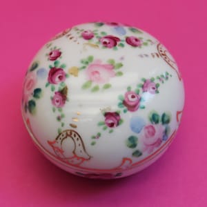 May include: A round white porcelain trinket box featuring hand-painted pink and dark red rose clusters with green leaves. The surface is decorated with gold-colored accents and thin orange lines along the base, creating a floral pattern.