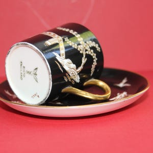 May include: Black teacup and saucer set with gold detailing and a white rim. The cup features a gold and white floral design and a gold handle. The saucer has a matching design with white bird silhouettes.