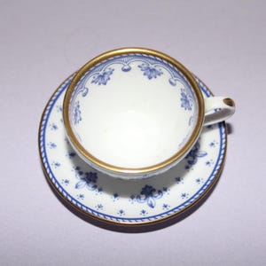 Puede incluir: Un juego de taza y platillo blancos con diseños florales azules y ribetes dorados. La taza tiene un asa delicada y el platillo tiene un diseño a juego. El juego es probablemente de porcelana y adecuado para servir té.