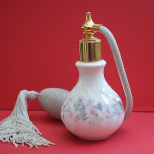 May include: A vintage white perfume bottle with a gold-colored top and a decorative floral pattern. The bottle has a bulb atomizer with a gray knitted bulb and a white tassel. The background is a solid red.
