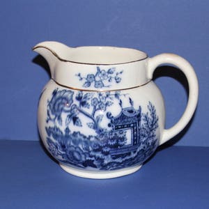 以下が含まれることがあります： 白のセラミッククリーマー、青と白の風景デザイン。クリーマーは丸みを帯びたボディ、湾曲したハンドル、注ぎ口を備えています。リムと注ぎ口は金でトリミングされています。デザインは、塔と花の要素が特徴です。